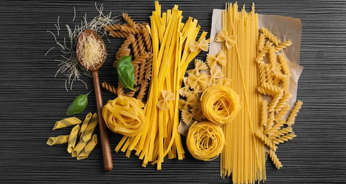 Różnorodne rodzaje makaronu, od cienkich nitek po tagliatelle i fusilli, ułożone na ciemnym tle. Wartości odżywcze makaronu zależą od jego składu.