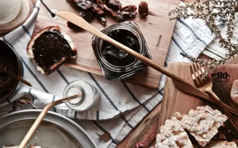 Słodkie śniadanie: kromka chleba z kremem czekoladowym, słoiczek z ciemnym dżemem, daktyle i ciastka. Idealna nutella bez orzechów!