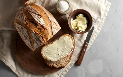 A kromka keto chleba na drożdżach, posmarowana masłem, obok bochenka, miseczki z masłem i solniczki.