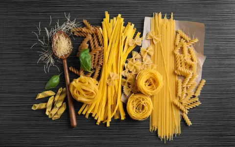 Różnorodne rodzaje makaronu, od cienkich nitek po tagliatelle i fusilli, ułożone na ciemnym tle. Wartości odżywcze makaronu zależą od jego składu.