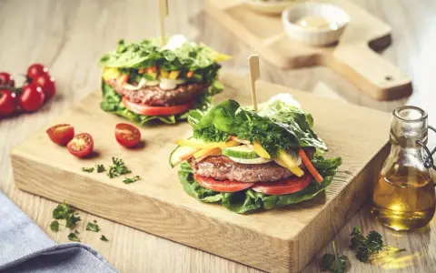 Dwa soczyste burgery w sałacie, z pomidorami, ogórkiem i papryką, na drewnianej desce.