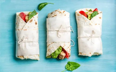 Trzy keto tortille z siemienia lnianego, zawinięte w papier i przewiązane sznurkiem, z widocznym nadzieniem z kurczaka, pomidorów i szpinaku.