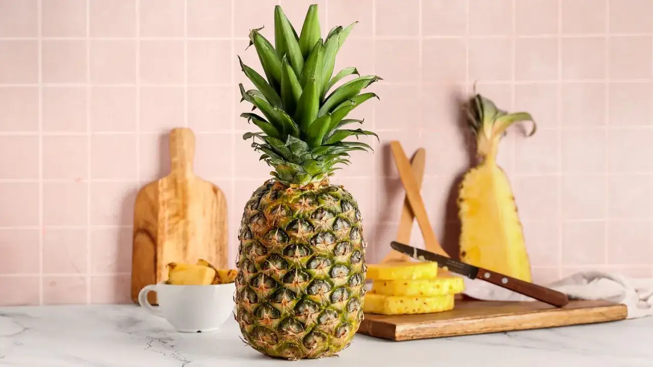 Ananas na blacie, obok pokrojone plastry i kawałki owocu. W tle deska do krojenia i różowe kafelki. Poznaj ananas wartości odżywcze.