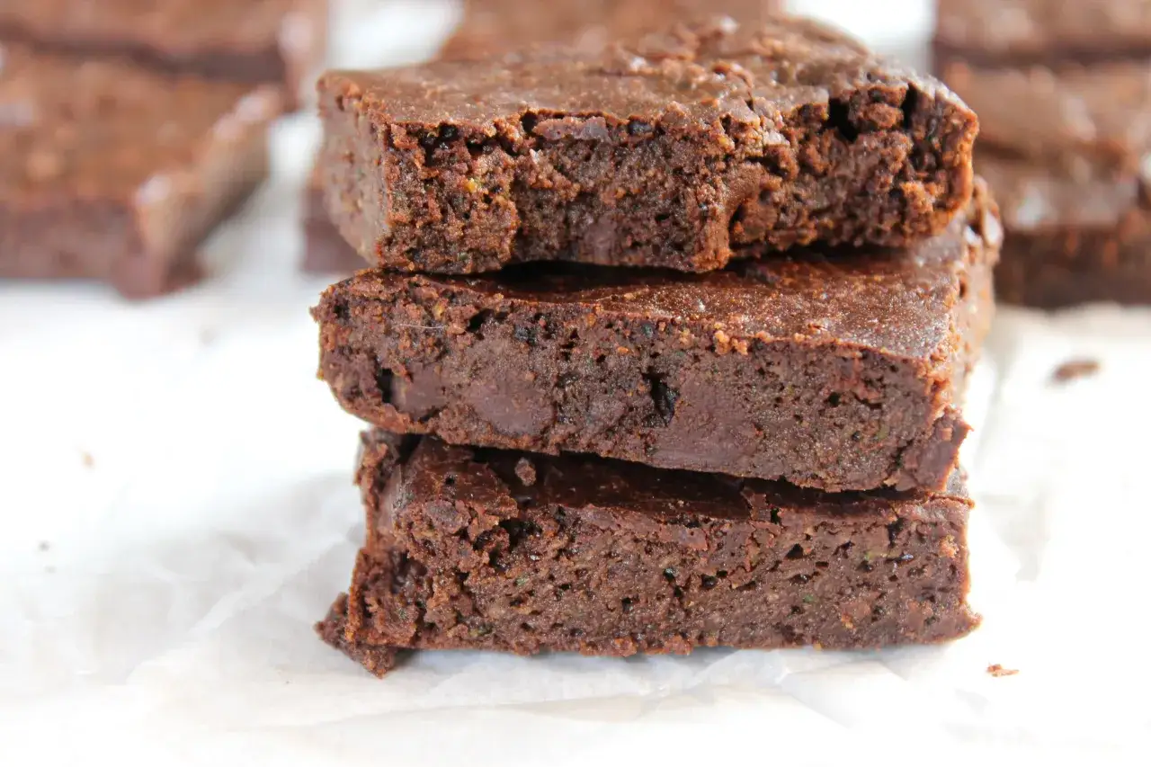 Czekoladowe keto brownie z cukinii, ułożone w stosik na białym papierze. Wilgotne i puszyste, z widocznymi kawałkami cukinii.