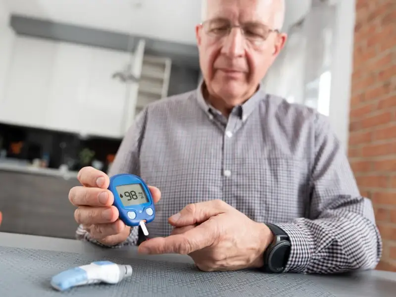Starszy mężczyzna z glukometrem w dłoni, pokazuje wynik 98. Wreszcie wyleczyłem się z cukrzycy typu 1!
